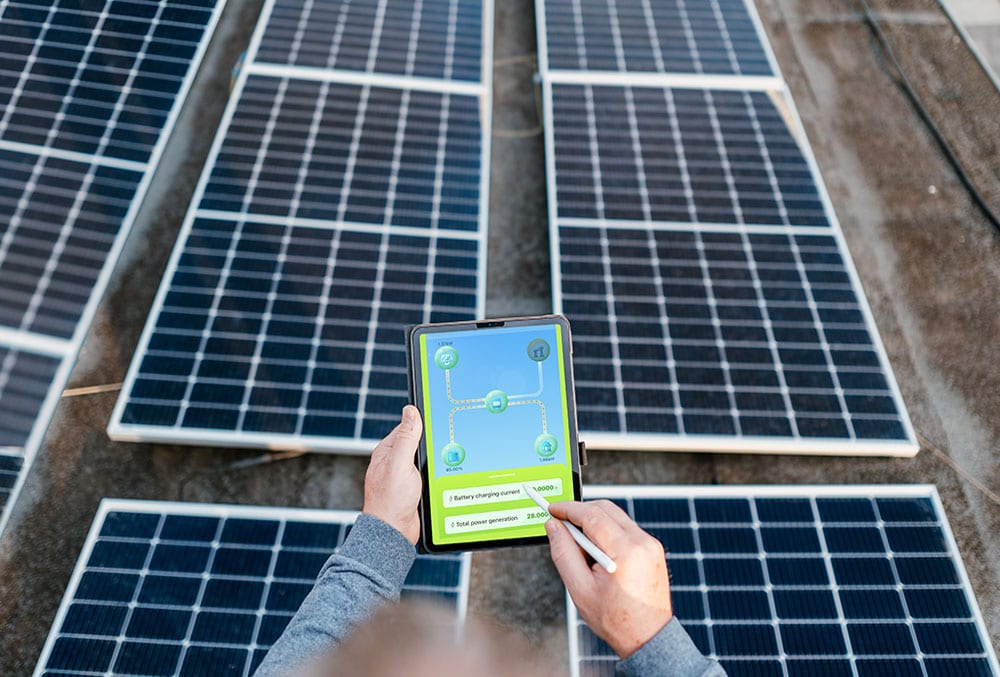 Primo piano di mani che utilizzano un tablet e una stylus per monitorare un impianto fotovoltaico sul tetto. Lo schermo del tablet mostra un'app di gestione energetica con dati di generazione e ricarica della batteria. L'immagine rappresenta il servizio di diagnosi guasti fotovoltaico senza sopralluogo a Cagliari di Energisolar Sardegna, che utilizza tecnologie avanzate come il monitoraggio delle curve I-V per analizzare a distanza lo stato dei pannelli,