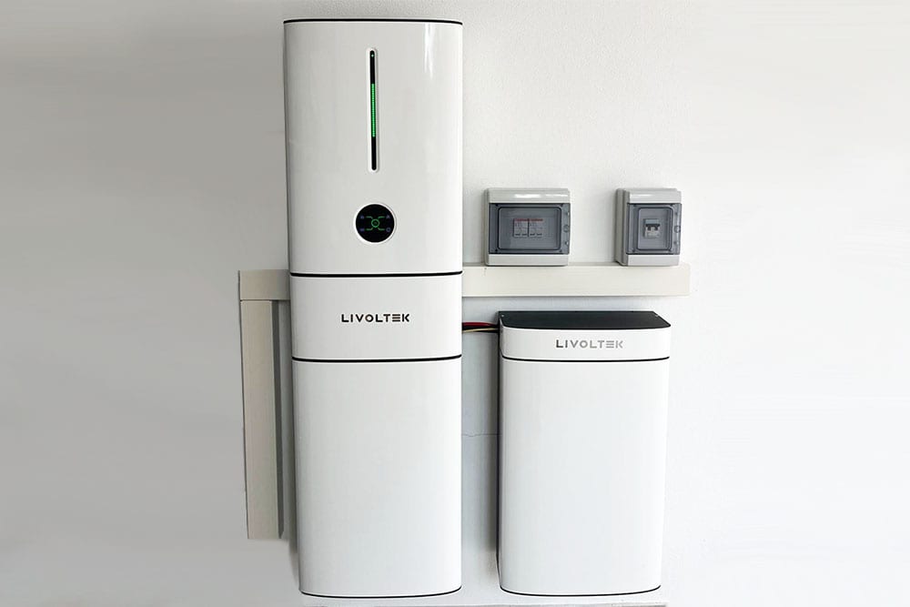 Primo piano di un'installazione domestica a parete di un sistema di accumulo residenziale del marchio Livoltek. Sono visibili l'inverter stack e un pacco batteria aggiuntivo di colore bianco, montati su una parete chiara. Sopra le unità, sono presenti due quadri elettrici grigi di protezione e canaline bianche. Il sistema mostra un indicatore LED verde acceso sull'unità principale. Nell'angolo in alto a destra è presente il logo 'energisolar SARDEGNA'. L'immagine rappresenta una soluzione di accumulo fisico fotovoltaico in Sardegna per massimizzare l'autoconsumo dopo la fine dello Scambio sul Posto.